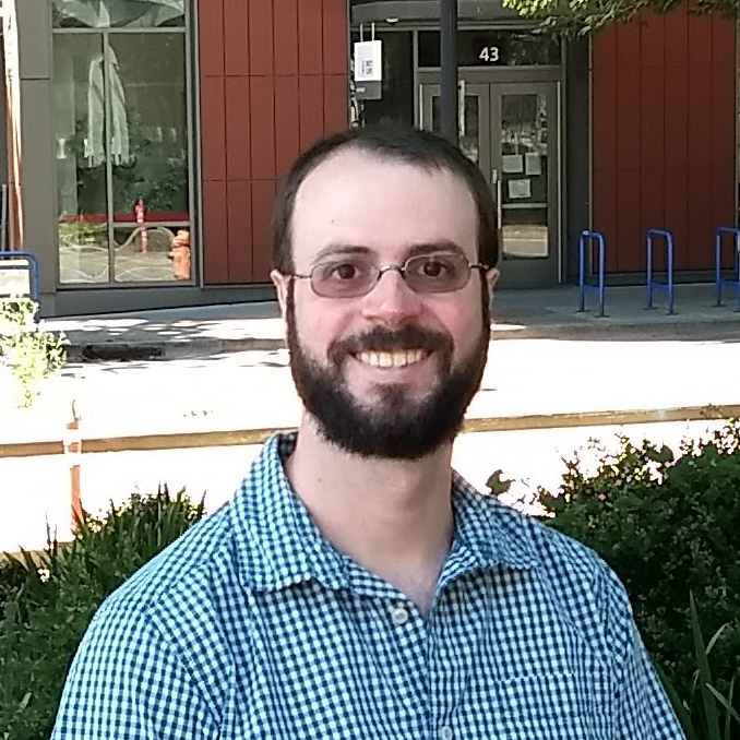 Photograph of a man with beard and glasses, seated outside in an urban setting. He is pictured from the shoulders up and is smiling.