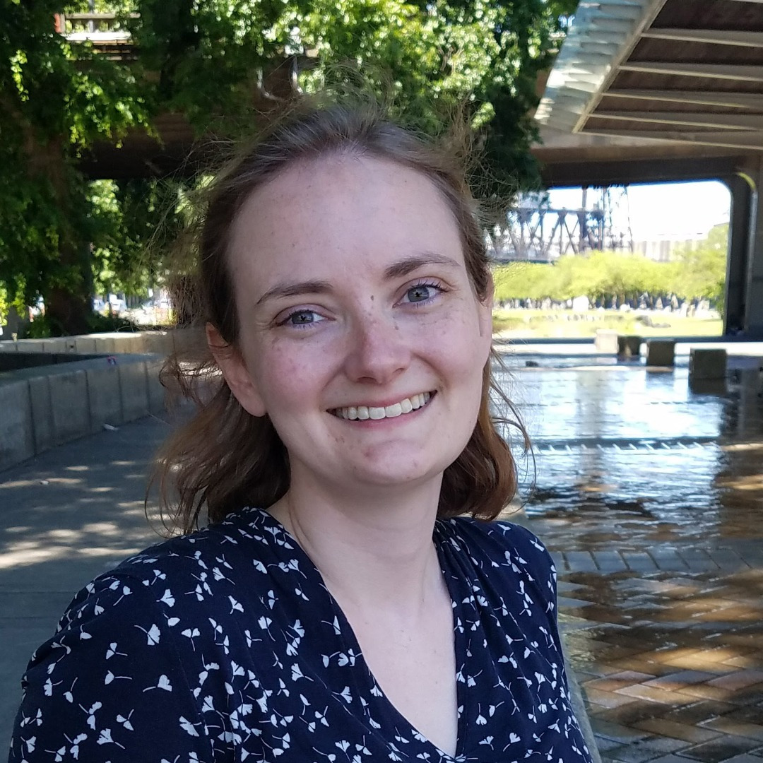 Photo of a woman smiling, pictured from the shoulders up. She sits outside in front of a fountain, with trees and bridge in the background.