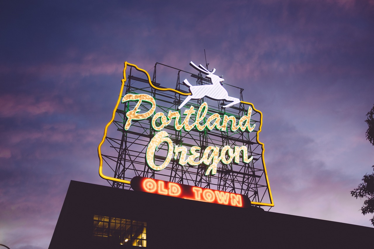 The Portland White Stag neon sign at dusk. The neon sign is lit up with an outline of the state of Oregon, a whit deer, ad the words "Portland, Oregon Old Town"